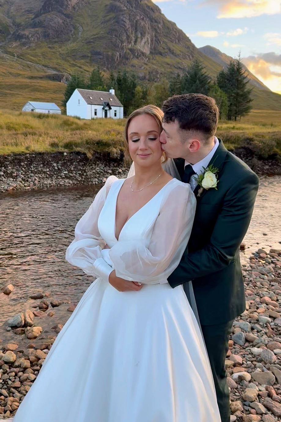 Grooms hugs bride from behind and kisses her cheek, with a small white house in the distance and hills in background.